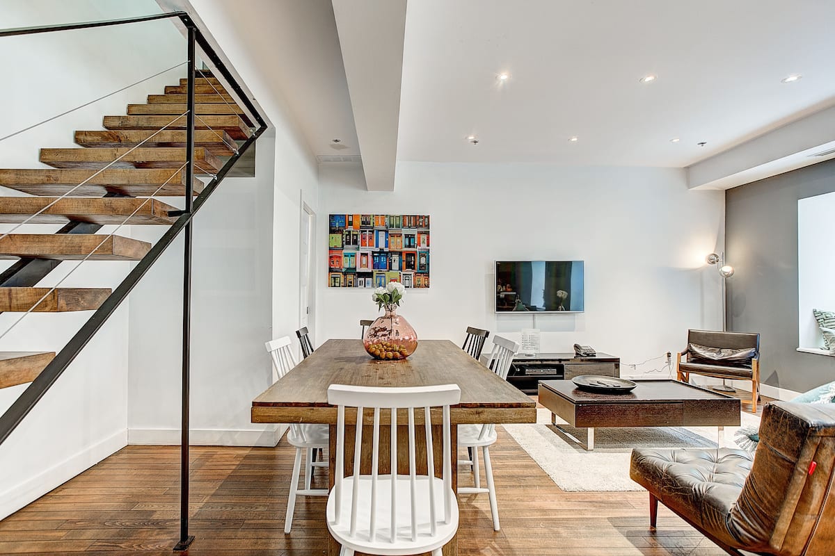 Una sala de estar moderna con una escalera de madera, una zona de comedor con una mesa y sillas de madera, un televisor de pantalla plana montado en la pared y un sofá de cuero.