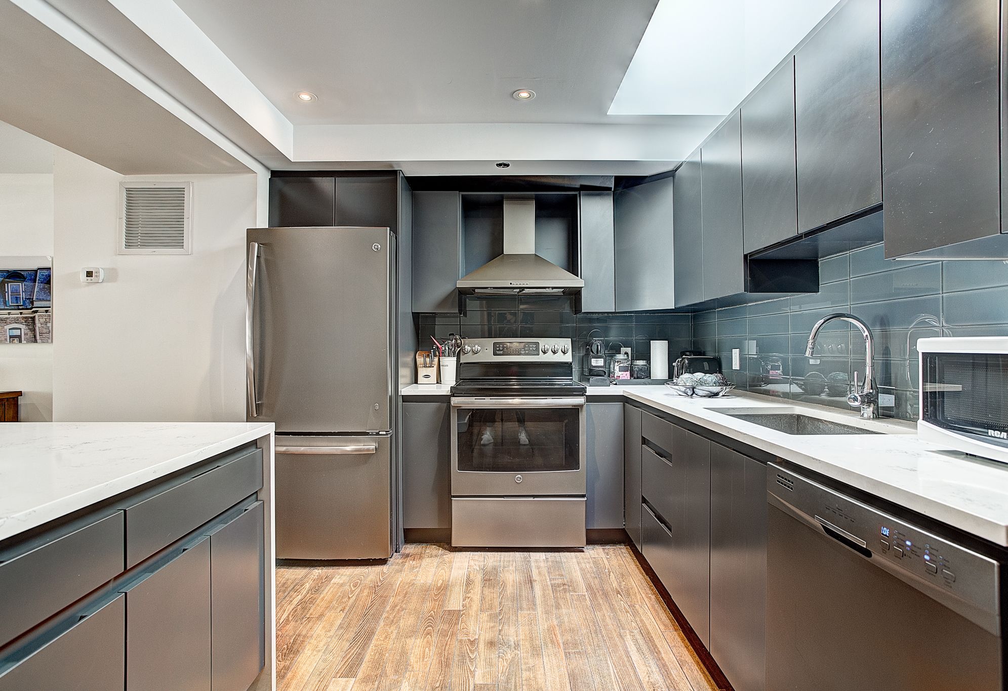 A modern kitchen with stainless steel appliances, grey cabinetry, and a wooden floor.