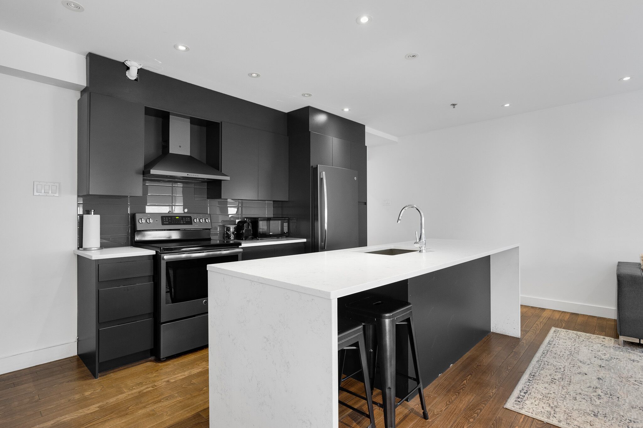 Une cuisine moderne avec des meubles de couleur sombre, des appareils en acier inoxydable, un îlot central doté de tabourets et un plancher en bois.