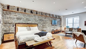 A bedroom with a stone wall, wooden floor, a bed with white linens, a bedside table, a nightstand, a TV mounted on the wall, and a window providing natural light. - The SFX Old Port (Montreal)