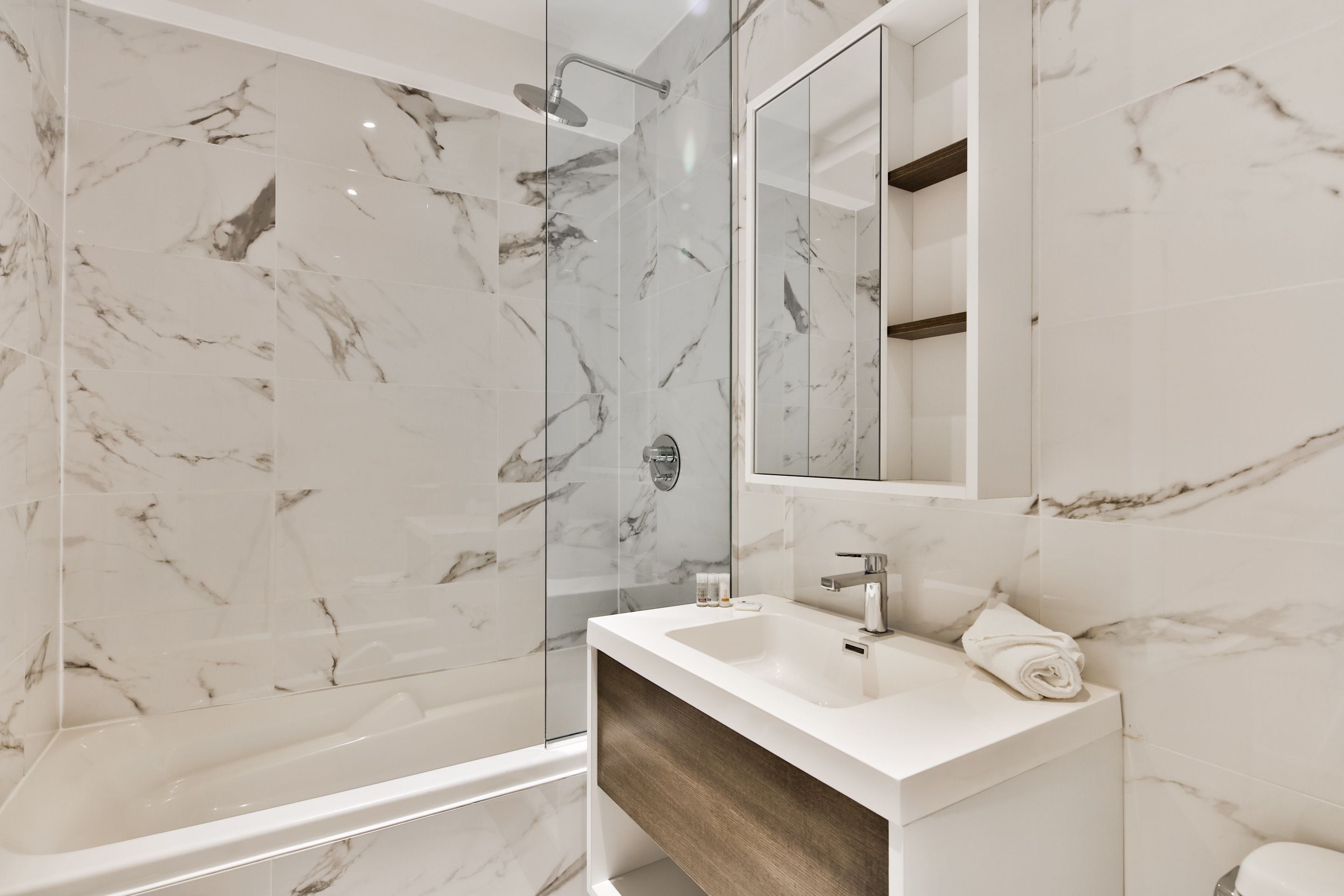 A modern bathroom with a marble shower, a white sink, and a wooden vanity.