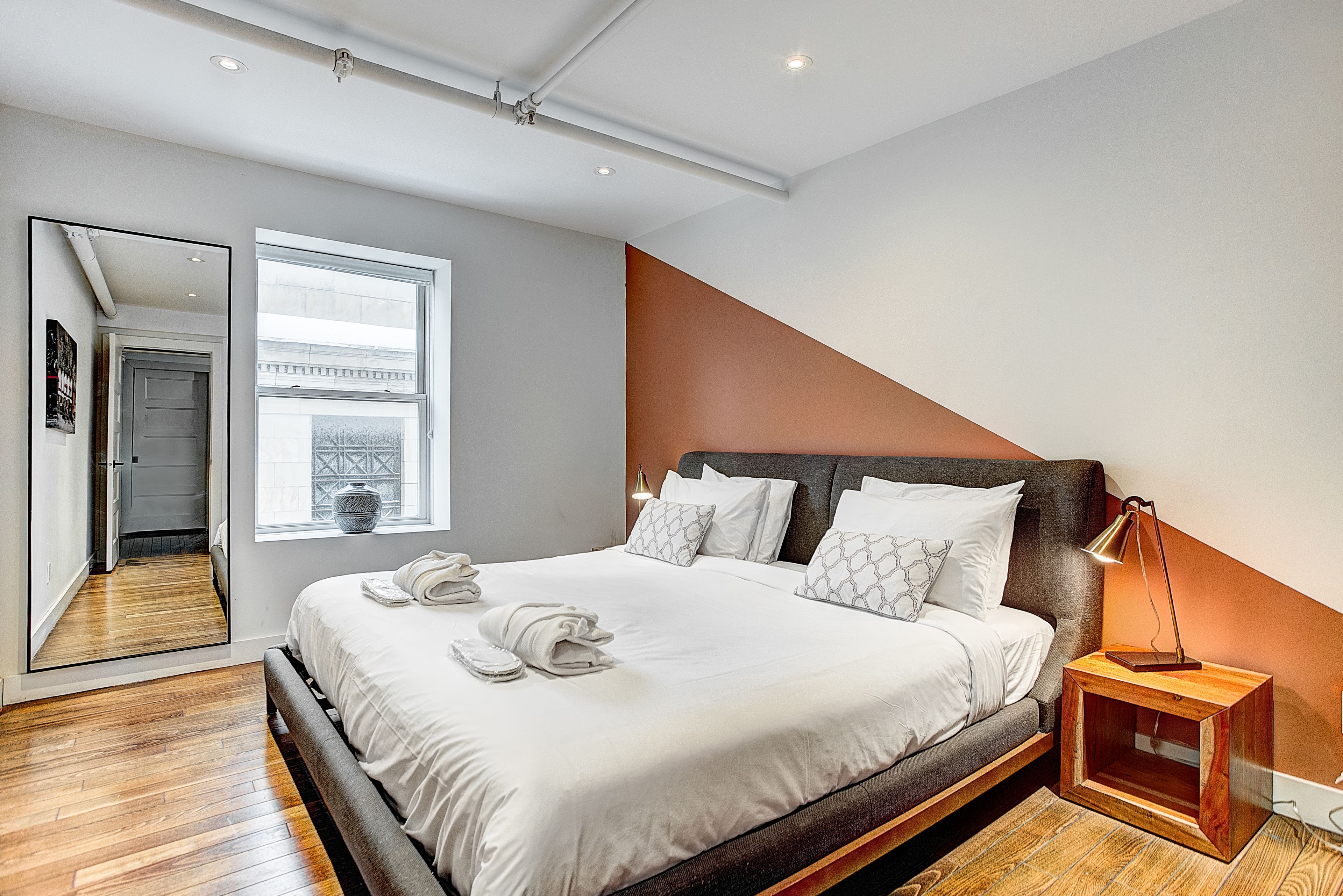 A modern bedroom with a large bed, a wooden floor, a window, and a nightstand with a lamp.