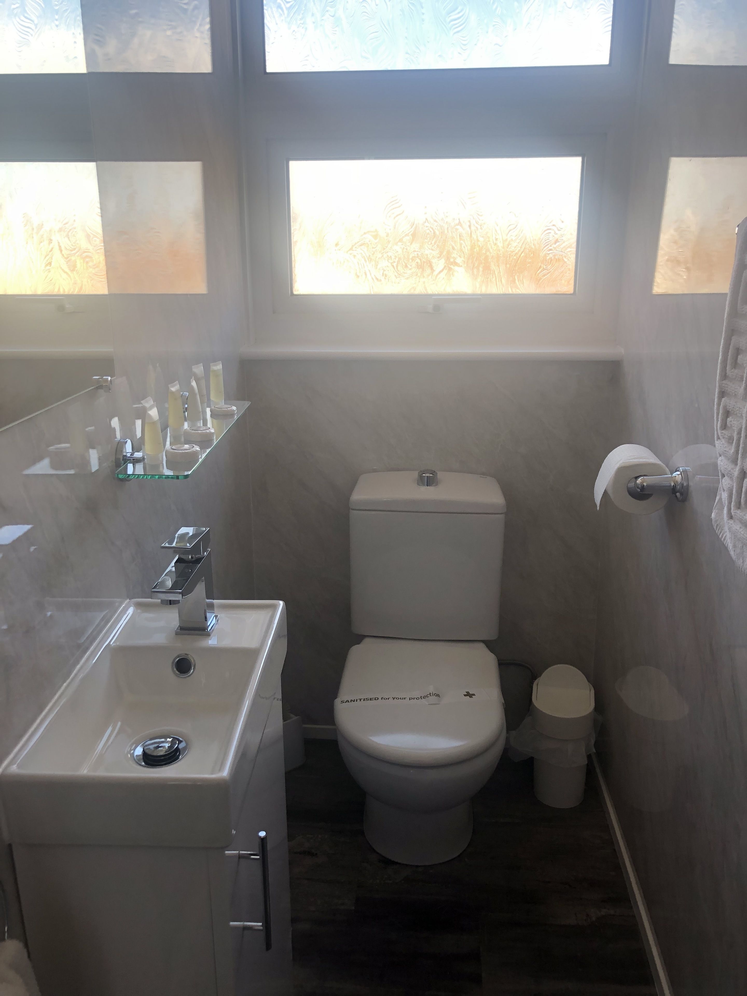 A compact bathroom with a toilet, sink, and glass shelf with toiletries.