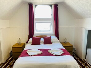 A neatly made bed with white and red bedding, two bedside lamps, and a window with curtains. - The Grosvenor House (Clacton-on-Sea)