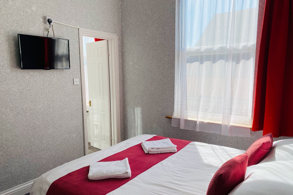 A hotel room with a bed, a television mounted on the wall, a door, and a window with sheer curtains.