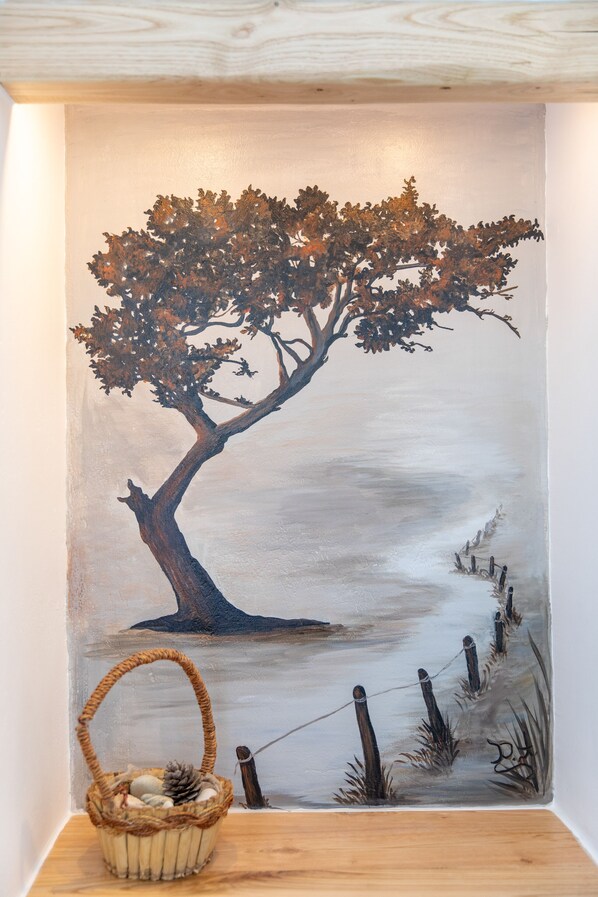 A wall with a painting of a tree by a body of water, a wooden bench, and a basket with stones. - Arhontiko Studios (Naxos)