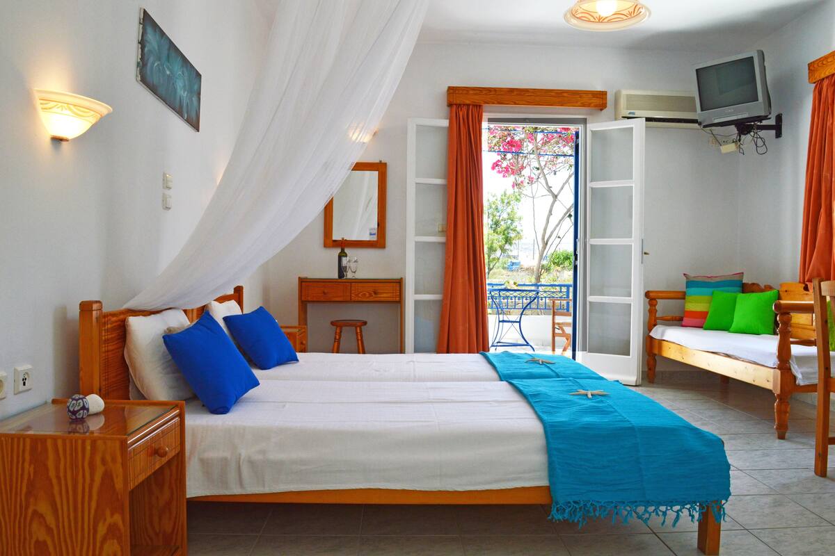A bedroom with a bed, a desk, a chair, a TV, and a balcony with a table and chairs.