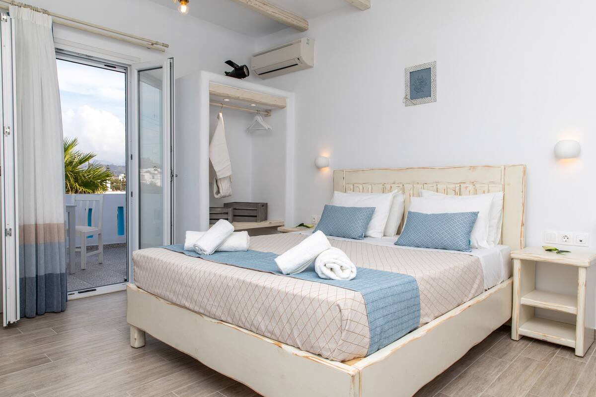 A bedroom with a bed, a nightstand, a wall-mounted light, and a view of the outside.