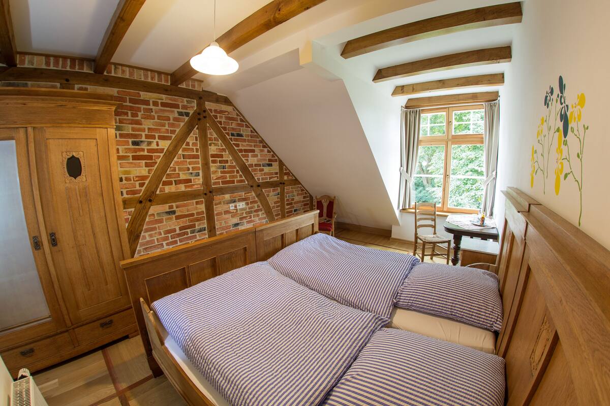 A bedroom with a sloped ceiling, exposed wooden beams, a brick wall, and a window with curtains.