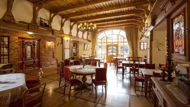 A spacious dining hall with high wooden ceilings, brick walls, and large windows allowing natural light. Tables are set with red chairs, and a chandelier hangs from the ceiling. - Landhaus Schulze Osthoff (Warendorf)