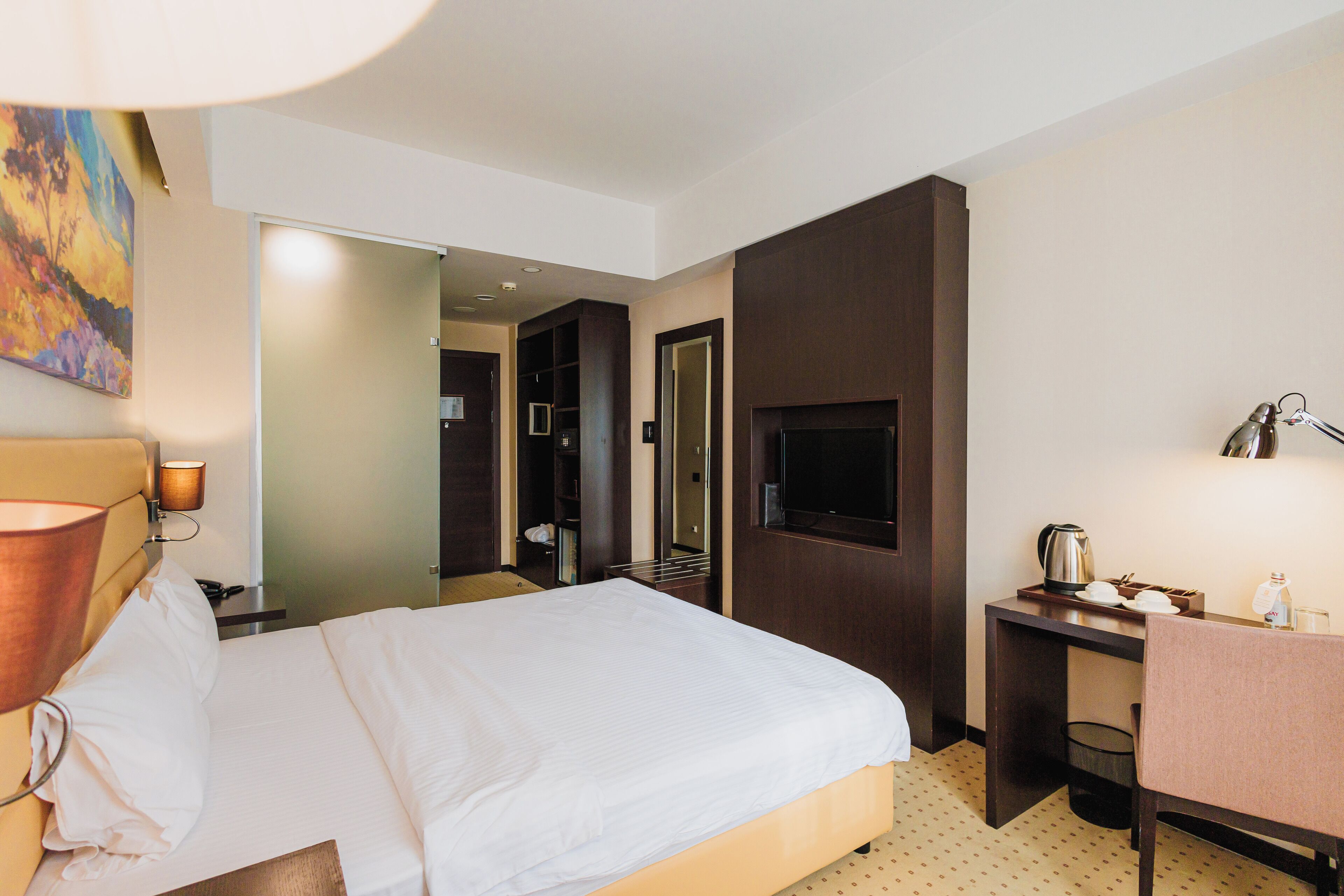 A hotel room with a bed, a desk with a lamp, a television, and a painting on the wall.