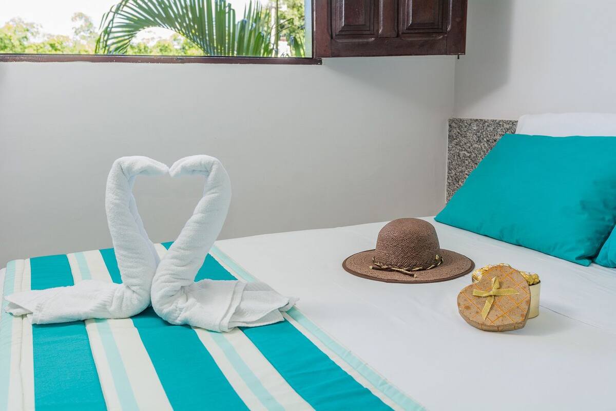 Une chambre d’hôtel avec un lit agrémenté de draps à rayures, une serviette pliée en forme de cœur, un chapeau de paille et une boîte cadeau en forme de cœur.