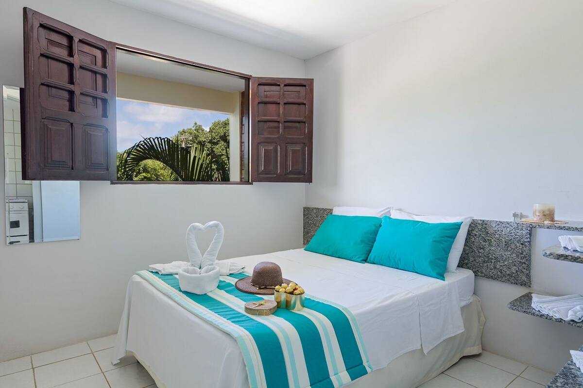 Une chambre d’hôtel avec un lit, un grand miroir, une fenêtre donnant sur un espace verdoyant et une petite étagère avec des serviettes.