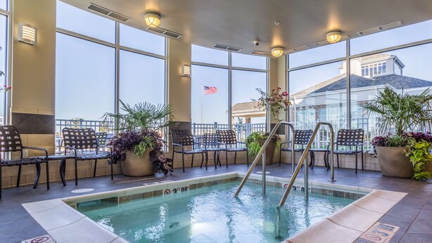 An indoor swimming pool with a ladder, surrounded by chairs and potted plants, large windows, and an American flag. - Hilton Garden Inn Salina (Salina)