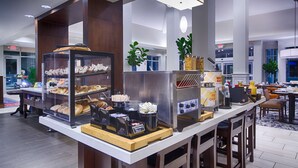 A hotel breakfast area with a self-service station, including a coffee machine and a variety of pastries displayed in a glass case. - Hilton Garden Inn Salina (Salina)