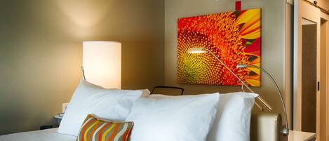 A hotel room with a bed, a bedside lamp, a colorful painting on the wall, and a door.