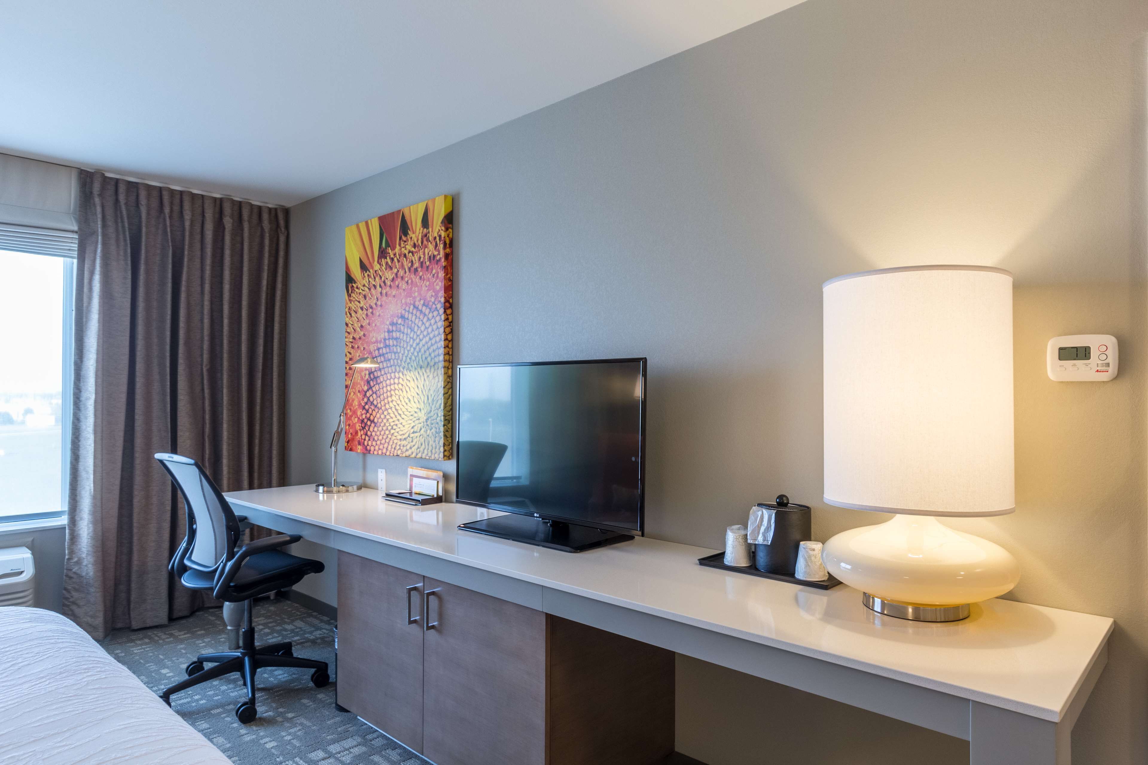 A hotel room with a bed, a desk with a TV, a lamp, and a thermostat on the wall.