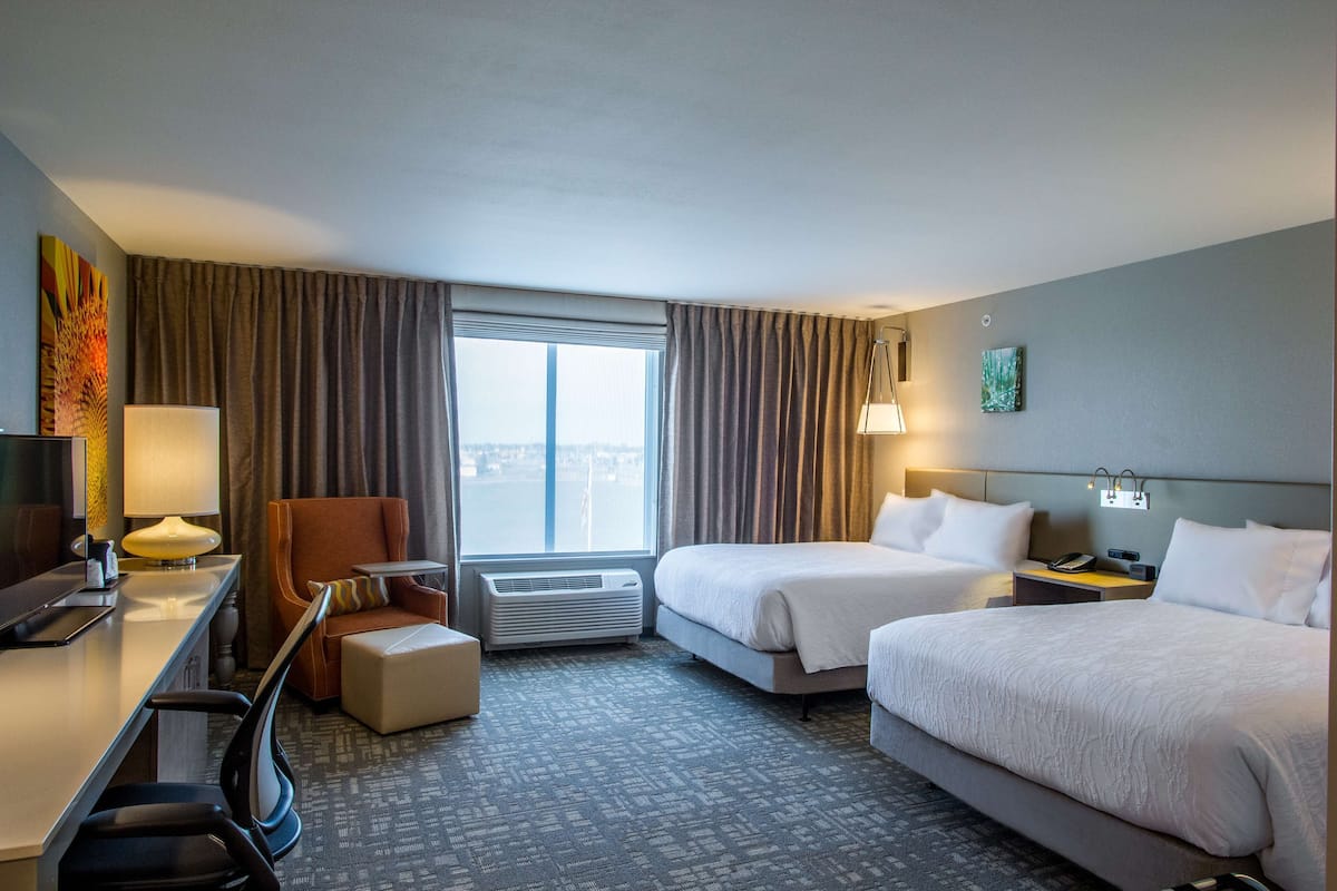 Une chambre d’hôtel avec deux lits, un bureau avec une chaise, une petite table, une lampe et une vue sur la ville grâce à de grandes fenêtres.