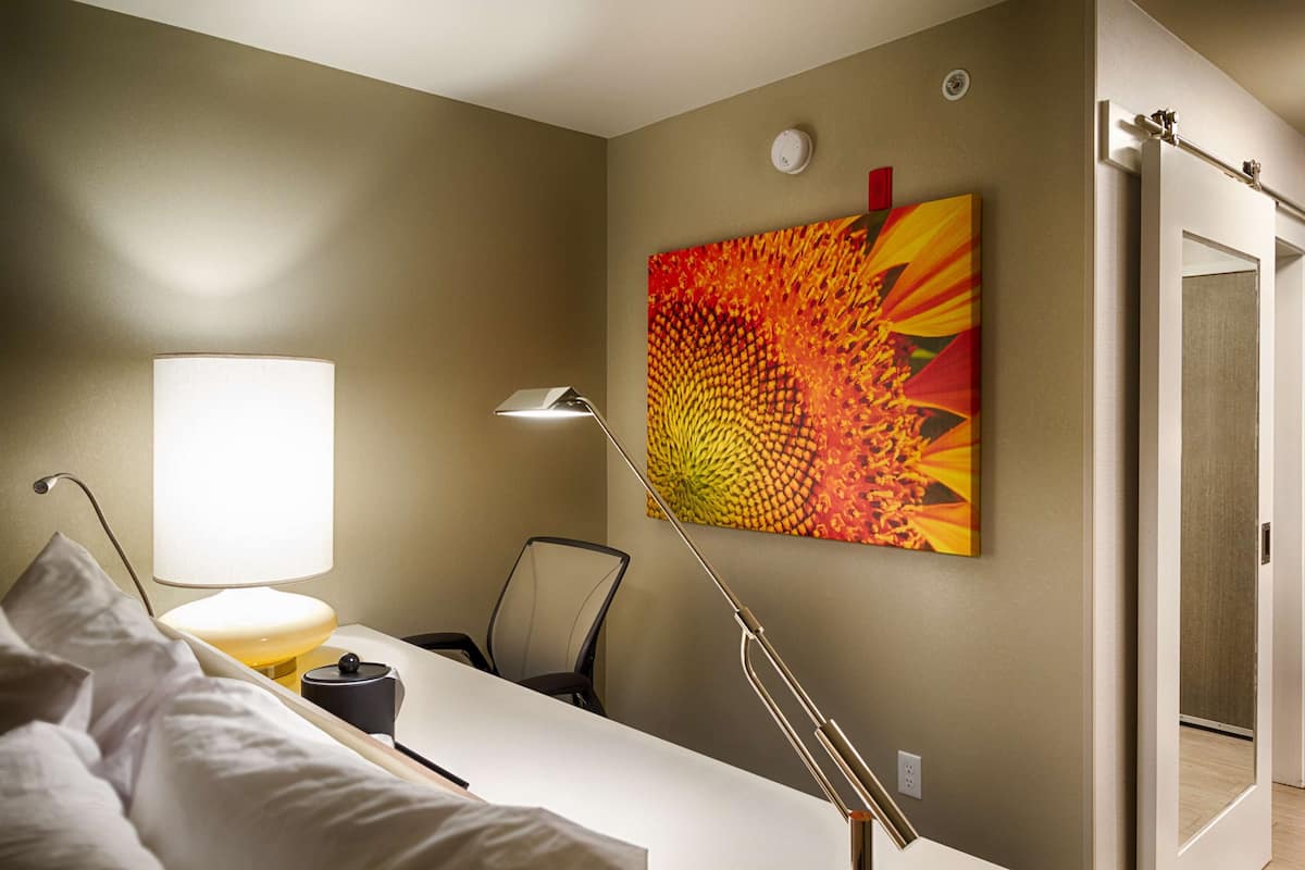 Une chambre d’hôtel avec un lit, un bureau, une chaise, une lampe et un grand tableau abstrait accroché au mur.