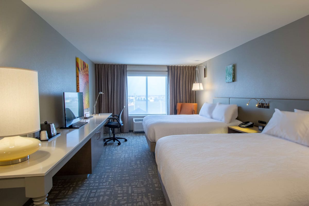 Une chambre d’hôtel avec deux lits, un bureau équipé d’un ordinateur et d’une lampe, et une vue sur la ville par la fenêtre.