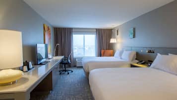 Quarto de hotel com duas camas, uma escrivaninha com computador, uma luminária e vista para a cidade pela janela.