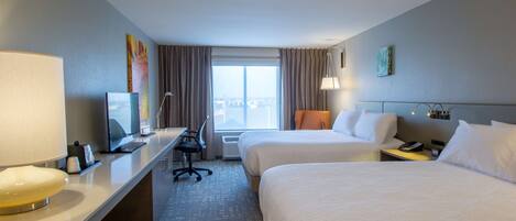 A hotel room with two beds, a desk with a computer, a lamp, and a view of the city through the window.