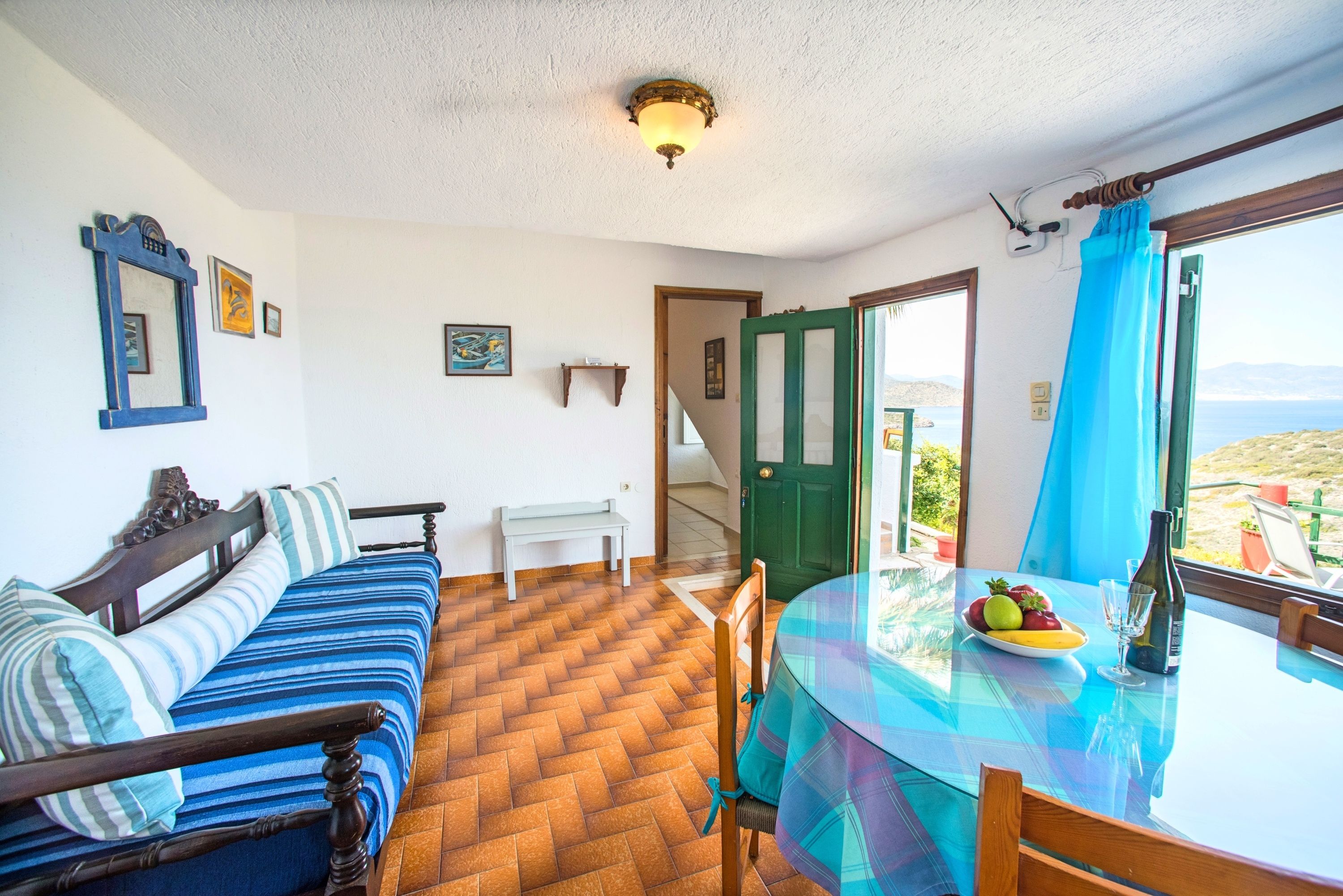 A room with a bed, a table with fruit, a view of the sea, and a green door.