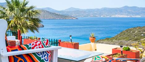 A terrace with a view of the sea and mountains, featuring a colorful chair and a table with potted plants.