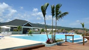 A resort pool area with a wooden deck, lounge chairs, and a building with a sloped roof. - Ocho Rios Getaway Villa at The Palms (Ocho Rios)