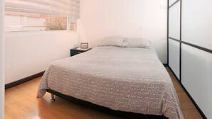 A bedroom with a bed, a nightstand, a window with blinds, and a view of the outside. - Studio Chapinero 216 (Bogotá)