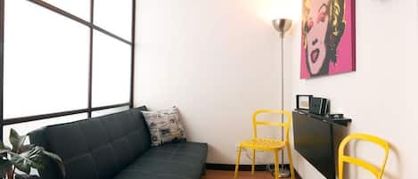 A modern living room with a black sofa, two yellow chairs, a small table, a floor lamp, a painting, and a wine rack.