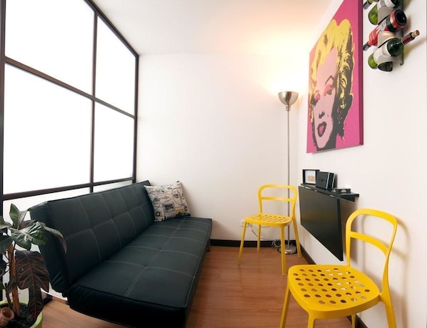 A modern living room with a black sofa, two yellow chairs, a small table, a floor lamp, a painting, and a wine rack.