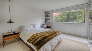 Quarto com uma janela grande, cama com manta de pele, mesa de cabeceira com abajur e estante de livros.