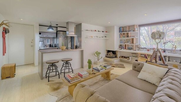 Uma sala de estar moderna com sofá, mesa de centro e estantes. Há uma área de cozinha com balcão e banquetas.