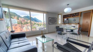 A modern living room with a black leather sofa, a glass coffee table, and a black and white chair. There is a dining area with a glass table and chairs, and a kitchen area with wooden cabinets and a hanging light fixture. - Apartment Zona Rosa 2204 (Bogotá)
