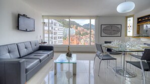 A modern living room with a sofa, a glass coffee table, a dining table set for four, and a large window offering a city view. - Apartment Zona Rosa 2204 (Bogotá)