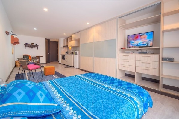 A modern bedroom with a large bed, a television, and a kitchenette in the background.