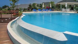 Ein Swimmingpool mit blauen Fliesen, umgeben von Liegestühlen und Sonnenschirmen. Im Hintergrund sind Palmen und ein Pavillon zu sehen.