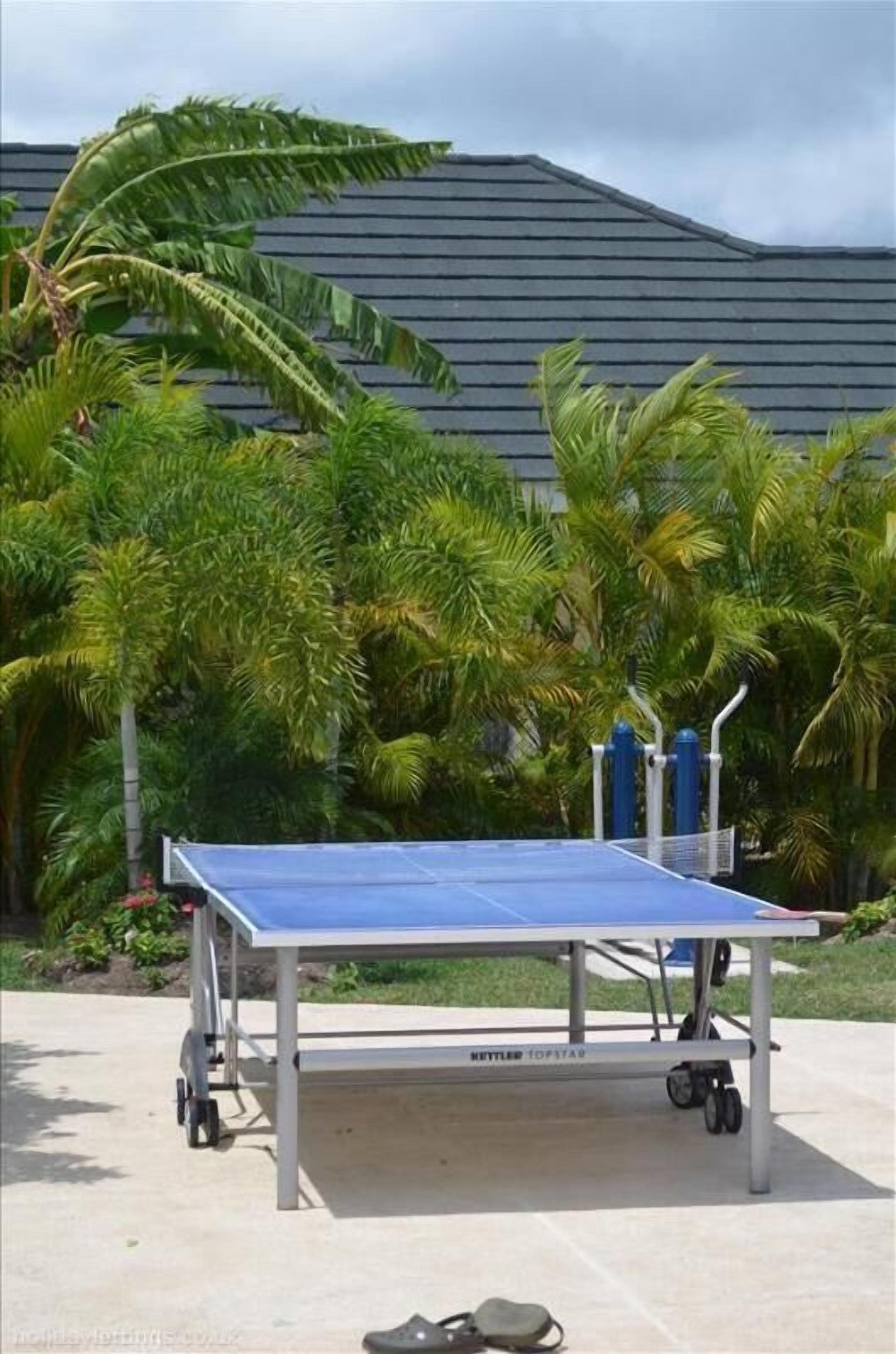 A ping pong table with a metal frame and wheels, set outdoors with palm trees and a building in the background.