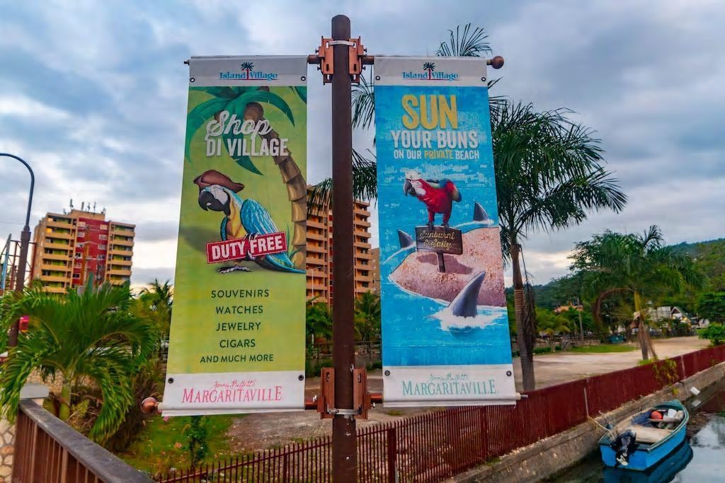 Two banners advertising Island Village with slogans "Shop DI VILLAGE DUTY FREE" and "SUN YOUR BUNS ON OUR PRIVATE BEACH".