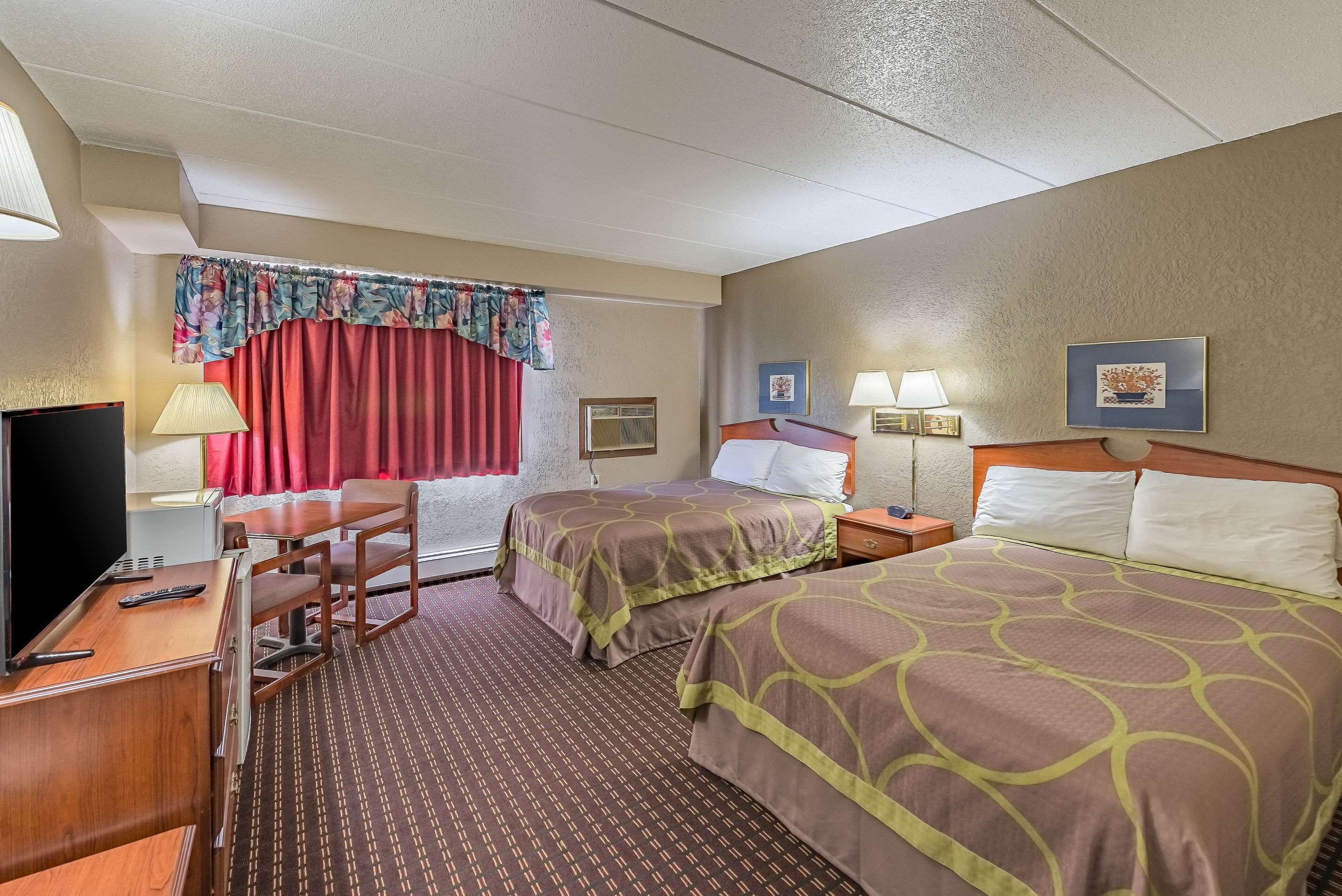 Habitación de hotel con dos camas, un escritorio con televisor, una silla, un ventanal con cortinas rojas y una pared con dos cuadros enmarcados.