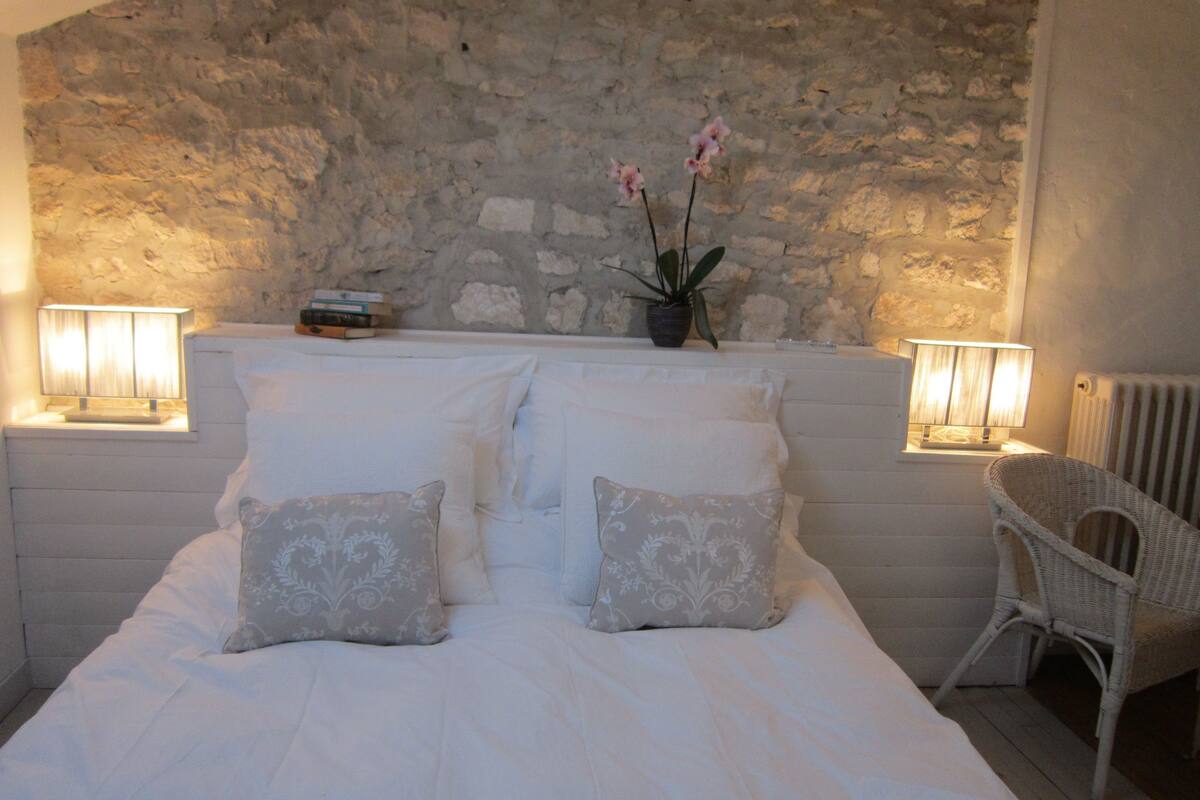 Une chambre avec un mur en pierre, un lit recouvert de linge de lit blanc, deux lampes sur les tables de chevet, une chaise en osier et une petite plante sur la table de chevet.