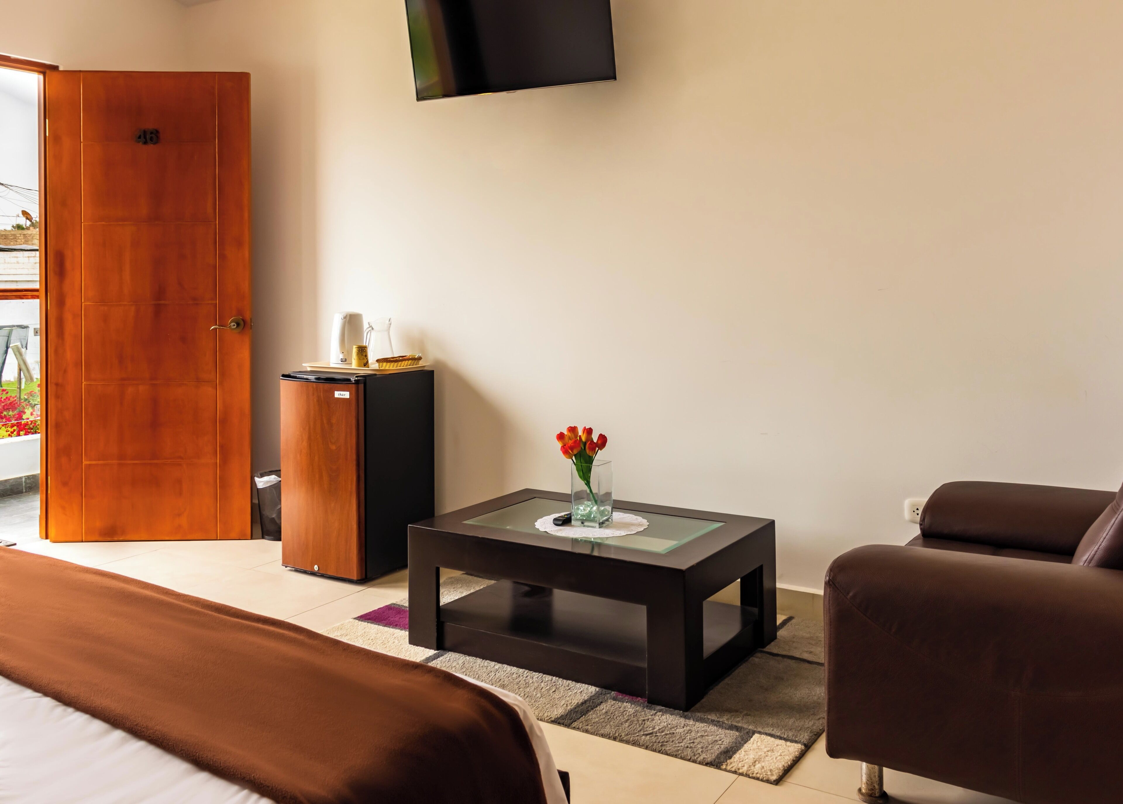 Een hotelkamer met een bed, een tafeltje met een bloevas, een aan de muur bevestigde televisie en een deur naar een buitenruimte.