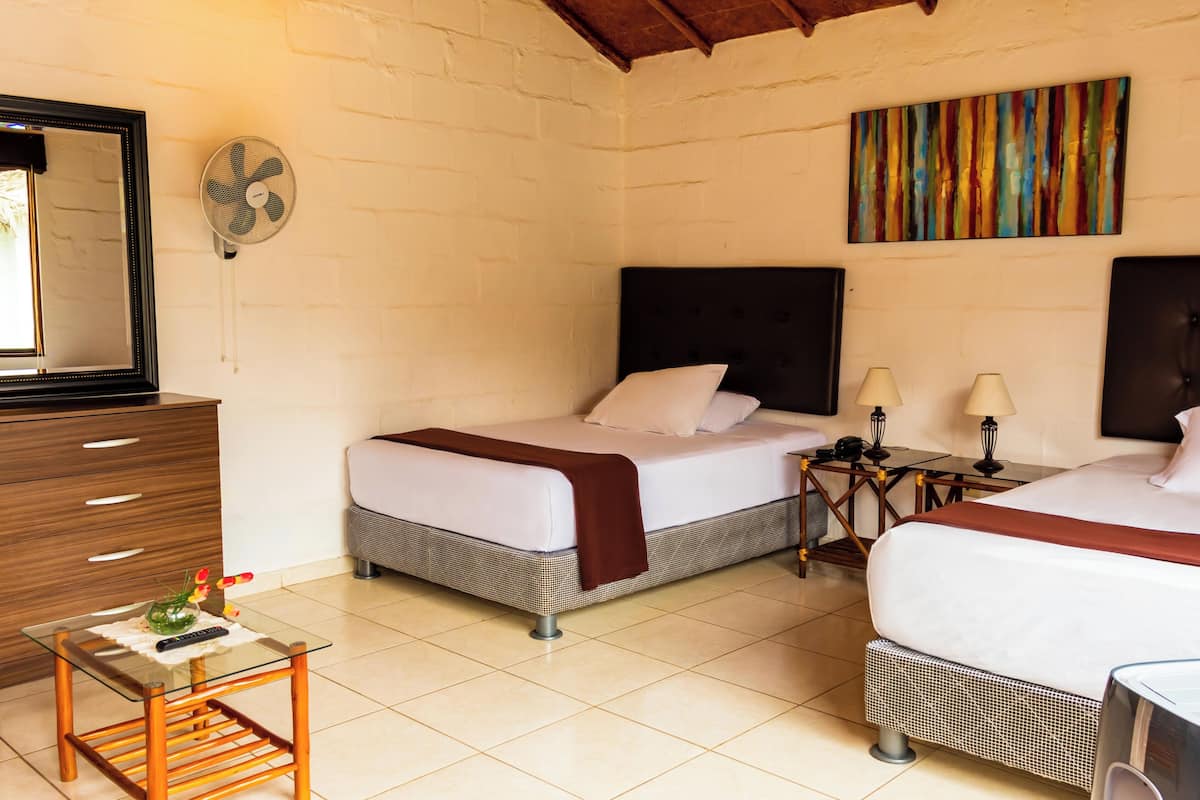 Ein Hotelzimmer mit zwei Betten, einem kleinen Tisch, einem Schminktisch und einem Ventilator. An der Wand hängt ein farbenfrohes abstraktes Gemälde.