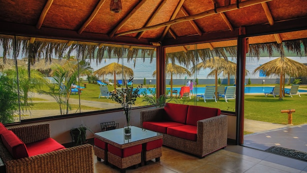 A covered outdoor seating area with wicker furniture and a glass table, overlooking a pool and beach. - QALA Hotel & Resorts (Grocio Prado)