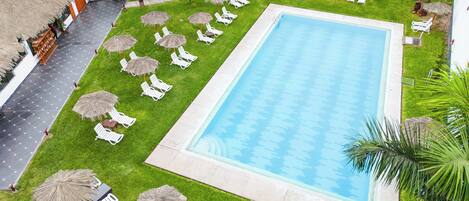 A swimming pool surrounded by white lounge chairs and thatched umbrellas, with a building in the background.