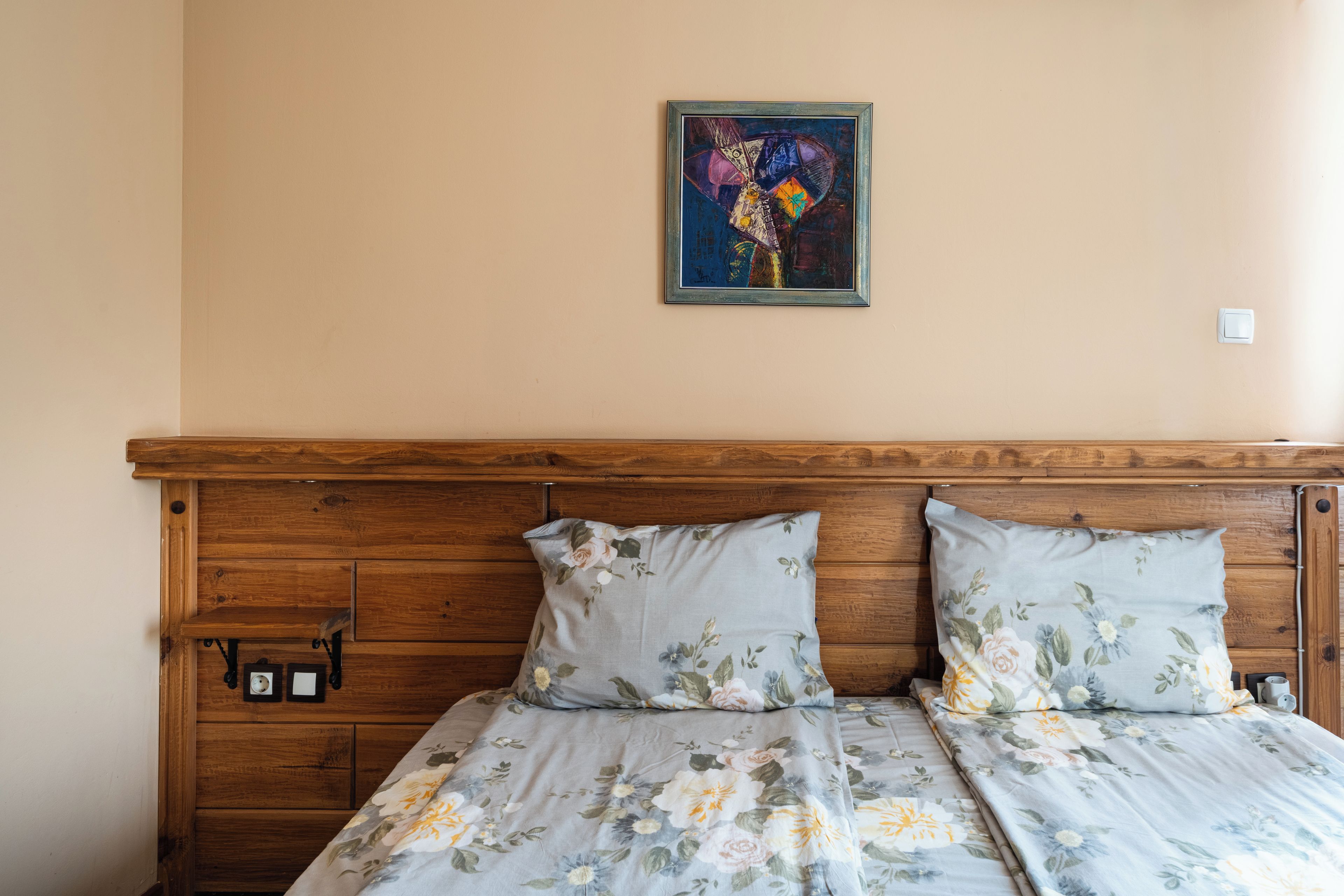 Una cama con ropa de cama floral, cabecero de madera, interruptor de luz en la pared y un cuadro abstracto enmarcado sobre la cama.