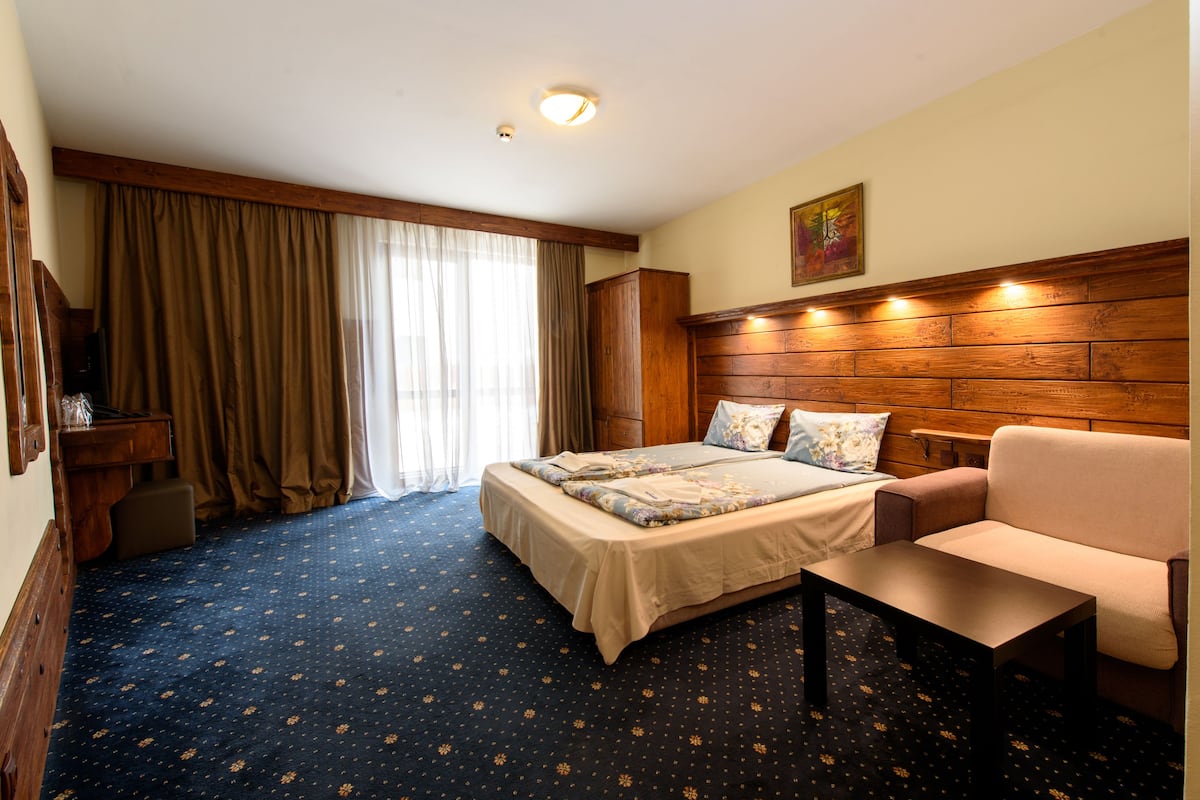 A hotel room with a bed, a sofa, a small table, a wooden headboard, and a window with curtains.