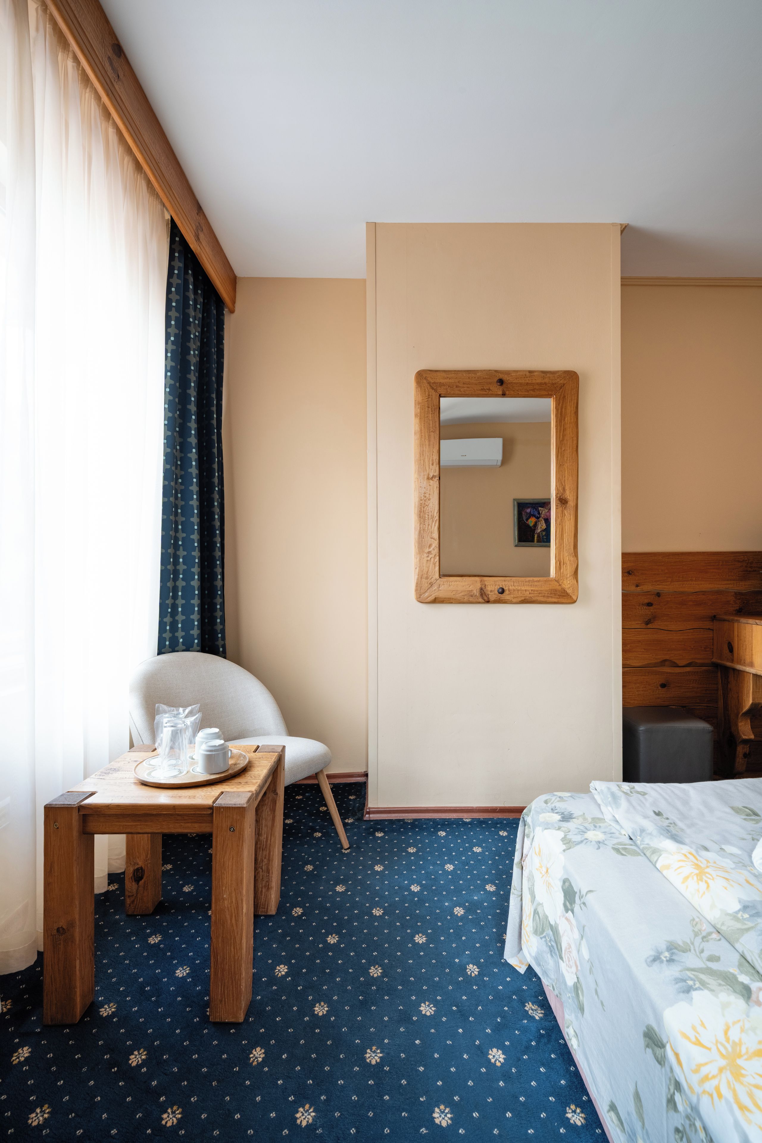 A hotel room with a wooden bed, a small table with cups, a chair, a mirror on the wall, and a patterned carpet.