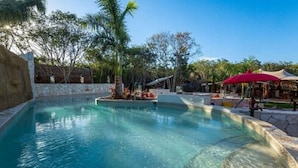 A clear swimming pool with a stone border, surrounded by palm trees and a shaded area with a red umbrella. - Serenity Authentic Glamping Tulum (Xpu-Ha)